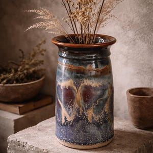 May include: A glazed ceramic vase in shades of blue, brown, and gold, holding dried grasses. The vase sits on a concrete block. A small wooden bowl and cup are in the background.