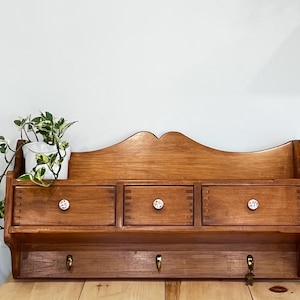 Peut inclure: Un organiseur mural en bois avec trois tiroirs et des crochets en laiton. Les tiroirs ont des boutons décoratifs. Un petit pot blanc avec une plante est posé dessus. Le bois a une finition chaude et naturelle.