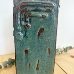 May include: A rectangular ceramic vase with a teal glaze and brown accents. The vase has raised, abstract designs on the front and top, with a textured surface. This decorative vase is a home decor item.