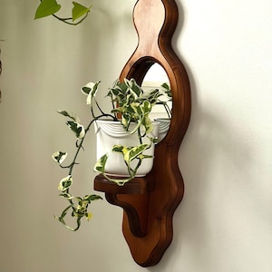 Op de afbeelding: Een houten wandplank met een ingebouwde spiegel en een klein plankje. Een witte keramische pot met een groene en gele plant staat op de plank. Het houten frame is donkerbruin en de spiegel is rond.