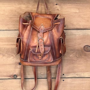 Op de afbeelding: Een bruine leren rugzak met een klepsluiting en verstelbare bandjes. De rugzak heeft twee zijzakken en een voorvak met een gesp. Het leer is gestikt met witte draad en de tas hangt aan een zwarte haak.