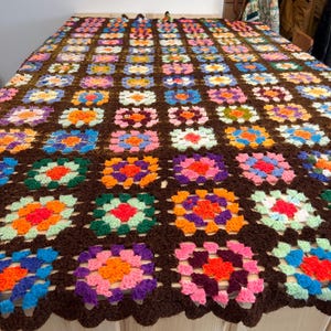 Op de afbeelding: Een handgemaakte gehaakte deken met een herhalend bloemenpatroon in levendige kleuren, waaronder roze, oranje, blauw en groen, tegen een donkerbruine achtergrond. De deken wordt getoond op een houten oppervlak, wat het ingewikkelde ontwerp en vakmanschap benadrukt.