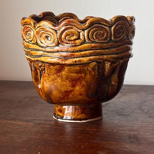 Può includere: Una ciotola in ceramica marrone con base a piedistallo. Il bordo presenta un motivo a spirale ripetuto. La ciotola ha una finitura lucida con variazioni di colore, che vanno dal marrone chiaro allo scuro. La ciotola è probabilmente fatta a mano.
