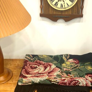 May include: A black textile with a large floral design in shades of pink, green, and yellow. The textile has a fringe along the bottom edge. A wooden clock with a cream-colored face and Roman numerals is in the background.