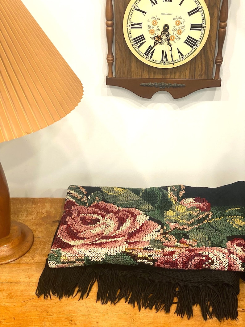 May include: A black textile with a large floral design in shades of pink, green, and yellow. The textile has a fringe along the bottom edge. A wooden clock with a cream-colored face and Roman numerals is in the background.