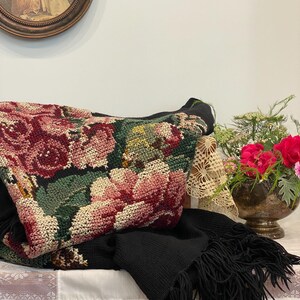 May include: A black knitted scarf with fringe is draped over a table, alongside a floral tapestry bag featuring pink and red roses. A vase of red flowers and a framed picture are also visible.