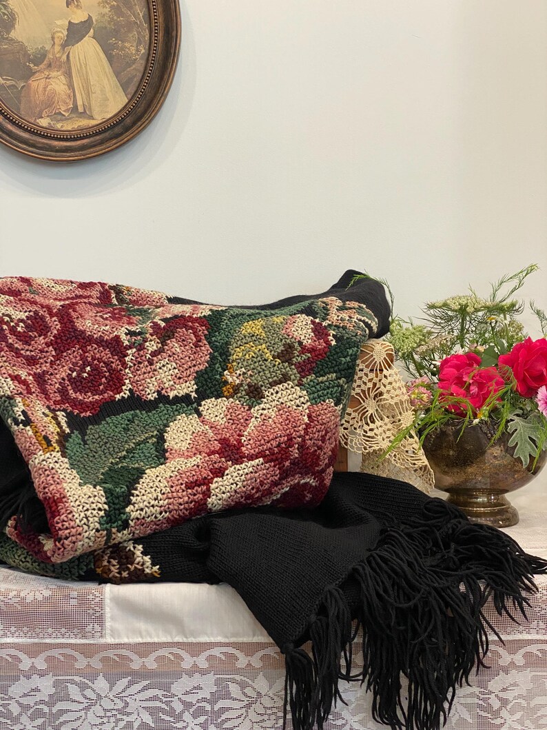 May include: A black knitted scarf with fringe is draped over a table, alongside a floral tapestry bag featuring pink and red roses. A vase of red flowers and a framed picture are also visible.