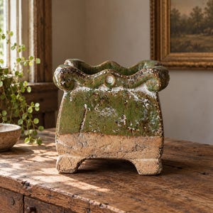 May include: A square ceramic planter with a scalloped rim and four short feet. The upper portion features a glossy, mottled green glaze, while the lower section displays a textured, unglazed beige clay finish with visible cracks.