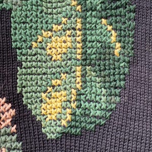 May include: Close-up of a dark blue knitted fabric with a detailed floral design. The design features green leaves with yellow accents and pink and red flowers, all created with cross-stitch embroidery.