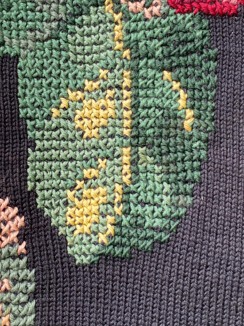 May include: Close-up of a dark blue knitted fabric with a detailed floral design. The design features green leaves with yellow accents and pink and red flowers, all created with cross-stitch embroidery.