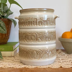 May include: A beige ceramic Rumtopf pot with a brown rim and handles. The pot features embossed fruit designs and the word "Rumtopf" in raised letters. The cylindrical pot is displayed on a wooden surface.