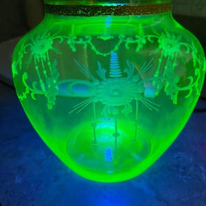 May include: A glowing green glass vase with etched floral designs. The vase has a wide, rounded body and a narrow neck with a gold-colored rim. The intricate patterns are illuminated, creating a striking visual effect. The vase is sitting on a dark surface.