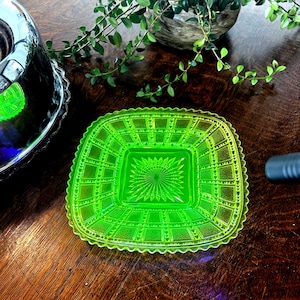 Può includere: Un piatto quadrato in vetro verde neon con un motivo strutturato e bordi smerlati. Il piatto ha un design centrale a forma di stella ed è appoggiato su una superficie di legno scuro. Un oggetto argentato e del verde sono visibili sullo sfondo.