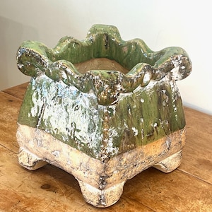May include: A square ceramic planter with a ruffled, scalloped rim. The upper portion features a glossy, deep green glaze with distressed white accents, while the lower base is a textured, unglazed tan clay with four sturdy feet.
