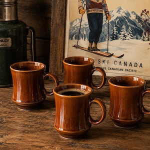 Può includere: Un set di quattro tazze in ceramica marrone con finitura lucida e una silhouette curva e affusolata. Ogni tazza presenta un manico robusto e un bordo liscio e arrotondato. Le tazze sono disposte in gruppo, con una tazza al centro in primo piano.