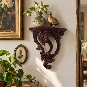 Può includere: Una mensola da parete marrone scuro con intagli ornamentali e dettagli floreali. La mensola sostiene un piccolo vaso in ceramica verde e bianco screziato con fiori bianchi e una statuetta di uccello in metallo dorato.