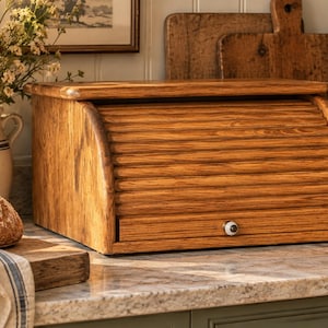 May include: A rectangular wooden bread bin with a curved, slatted roll-top door and a small, round white knob. The bin features a natural wood grain finish and is constructed with a flat top surface and a solid base, providing a storage solution.