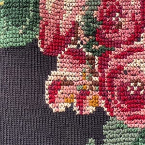 May include: Close-up of a cross-stitched floral design. The design features pink, red, and white flowers with green leaves and yellow accents. The background is a dark navy blue knit fabric.