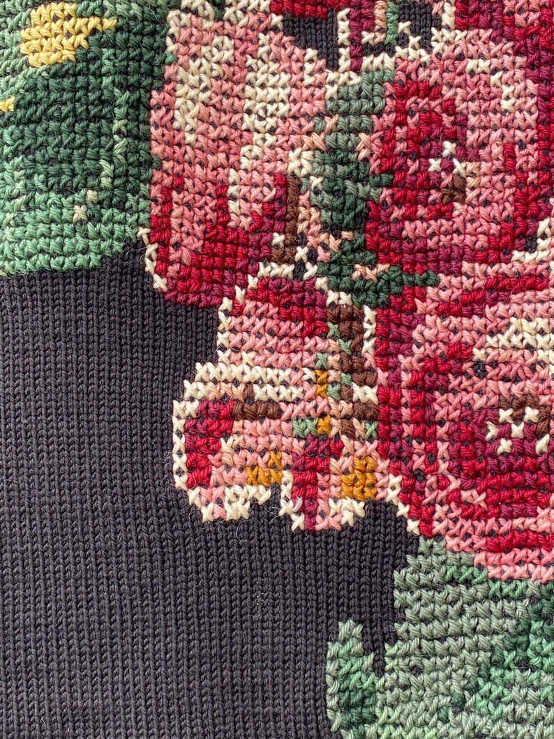 May include: Close-up of a cross-stitched floral design. The design features pink, red, and white flowers with green leaves and yellow accents. The background is a dark navy blue knit fabric.