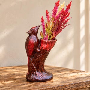 May include: A glossy dark red ceramic vase shaped like a bird perched on a tree trunk. The vase features a sculpted cardinal figure with a smooth finish and contains a bouquet of vibrant pink, red, and yellow dried celosia flower stems.