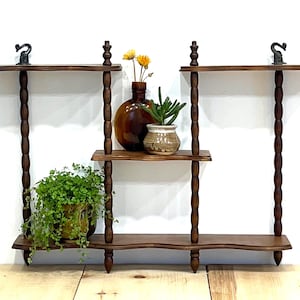 May include: A dark brown, three-tiered wooden wall shelf with decorative turned supports. The shelf is adorned with a variety of plants and decorative vases. The top shelves have decorative metal accents.