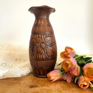 May include: A brown wooden vase with a carved floral design. The vase is sitting on a wooden table with a bouquet of orange and pink tulips.
