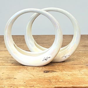 May include: Two white, iridescent ceramic ring-shaped vases. Each vase has a curved, open design with a flat base and a circular top. The vases are on a wooden surface. The text "Lady Grace" is written on the side of each vase.