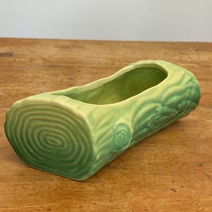May include: A light green ceramic planter shaped like a log. The planter has a hollowed-out interior and is decorated with tree ring and bark details. The planter is a decorative item for plants.