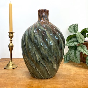 May include: A large, glazed ceramic vase with a swirling, textured design in shades of green, brown, and bronze. The vase has a wide, rounded body and a narrow neck. A brass candlestick with a lit, yellow candle is to the left.