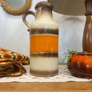 Może przedstawiać: Wazon ceramiczny w stylu vintage z uchwytem, w kolorach kremowym, pomarańczowym i brązowym. Cylindryczny wazon ma szeroką podstawę i wąską szyjkę, z poziomymi pasami koloru. Retro design.