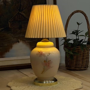 May include: A vintage table lamp with a pleated, cream-coloured lampshade. The base is a light pink ceramic urn with a floral design. The lamp sits on a gold-coloured base and a cream-coloured doily.