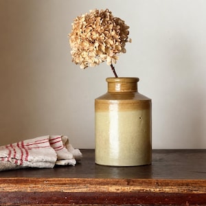 May include: A cylindrical stoneware crock with a two-tone glaze featuring a tan upper section and a light beige lower body. A single dried hydrangea flower with light brown petals is placed inside the crock. A folded white cloth with red stripes.