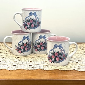 Könnte beinhalten: Vier weiße Keramikbecher mit rosa Innenraum und Henkel. Jeder Becher hat ein Design eines Blumenkorbs mit einer blauen Schleife und Tupfen. Die Becher sind auf einer dekorativen Spitzendecke angeordnet.