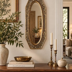 May include: An oval mirror with a gold-toned, ornate, hexagonal frame featuring intricate scrollwork and floral details. The mirror is mounted on a wall above a wooden console table, reflecting a room interior with a doorway and a framed picture.