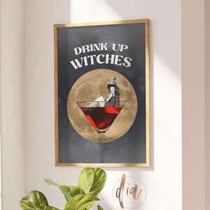 K&ouml;nnte beinhalten: Ein gerahmter Druck mit schwarzem Hintergrund und goldenem Rahmen. Der Druck zeigt eine Hexe, die in einem Martini-Glas sitzt, mit dem Text "Drink Up Witches" dar&uuml;ber.