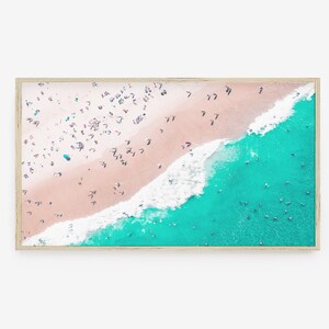 May include: An aerial view of a beach with turquoise water and white waves crashing on the shore. The sand is pink and there are many people on the beach.