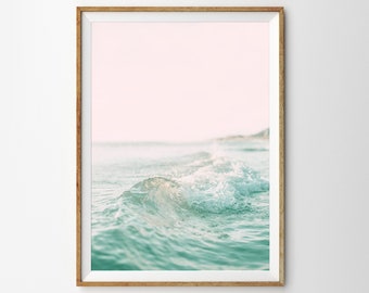 May include: A close-up photograph of a wave crashing on a beach. The water is a light blue-green color and the sky is a soft pink.