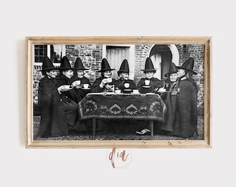 May include: A black and white photograph of a group of women dressed as witches, sitting around a table with teacups and a teapot. They are wearing black cloaks and pointed hats.