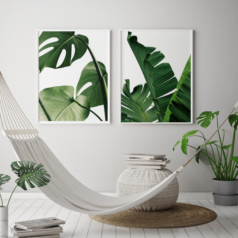 May include: Two framed prints of tropical green leaves, one with a monstera leaf and the other with a banana leaf, hanging on a white wall above a white hammock.