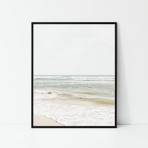 May include: A framed print featuring a serene beach scene. The image captures the ocean with gentle waves rolling onto a sandy shore. The sky is a soft, muted color, creating a calming coastal aesthetic.