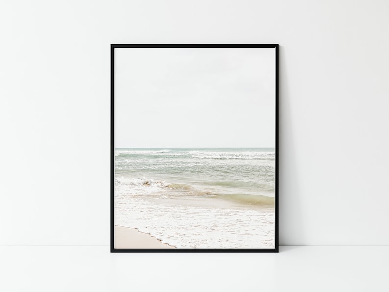May include: A framed print featuring a serene beach scene. The image captures the ocean with gentle waves rolling onto a sandy shore. The sky is a soft, muted color, creating a calming coastal aesthetic.