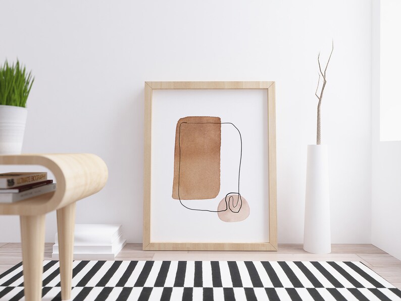 Op de afbeelding: Een ingelijst abstract kunstprint met een bruin en roze aquarelontwerp en een zwarte lijntekening. De print is in een licht houten lijst en hangt aan een witte muur.