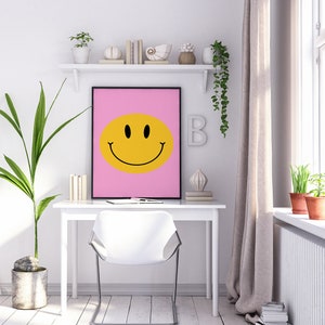 May include: A white desk with a white chair in front of it. A pink framed poster with a yellow smiley face on it hangs above the desk. There are plants and decorative items on a shelf above the desk.