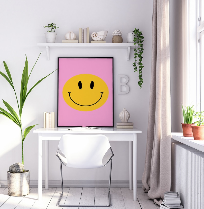 May include: A white desk with a white chair in front of it. A pink framed poster with a yellow smiley face on it hangs above the desk. There are plants and decorative items on a shelf above the desk.