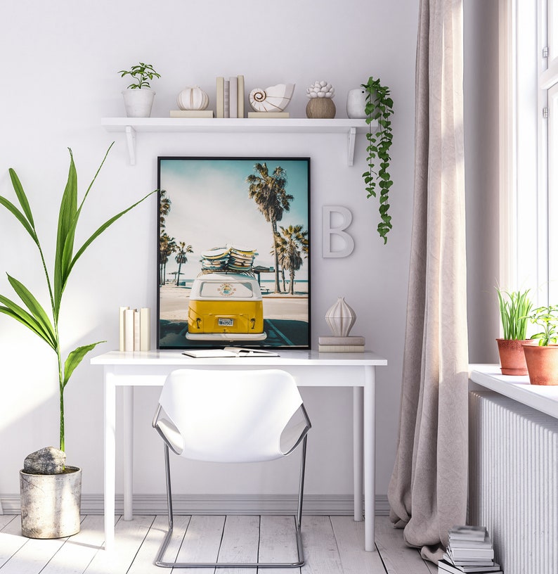 May include: A white desk with a white chair and a framed print of a yellow vintage van with surfboards on top, parked on a beach with palm trees in the background.