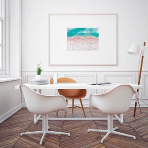 Op de afbeelding: Een wit bureau met een ingelijste foto van een luchtfoto van een strand met turquoise water en roze zand. Twee witte stoelen en een bruine stoel staan rond het bureau. Een lamp met een witte kap staat op de achtergrond.