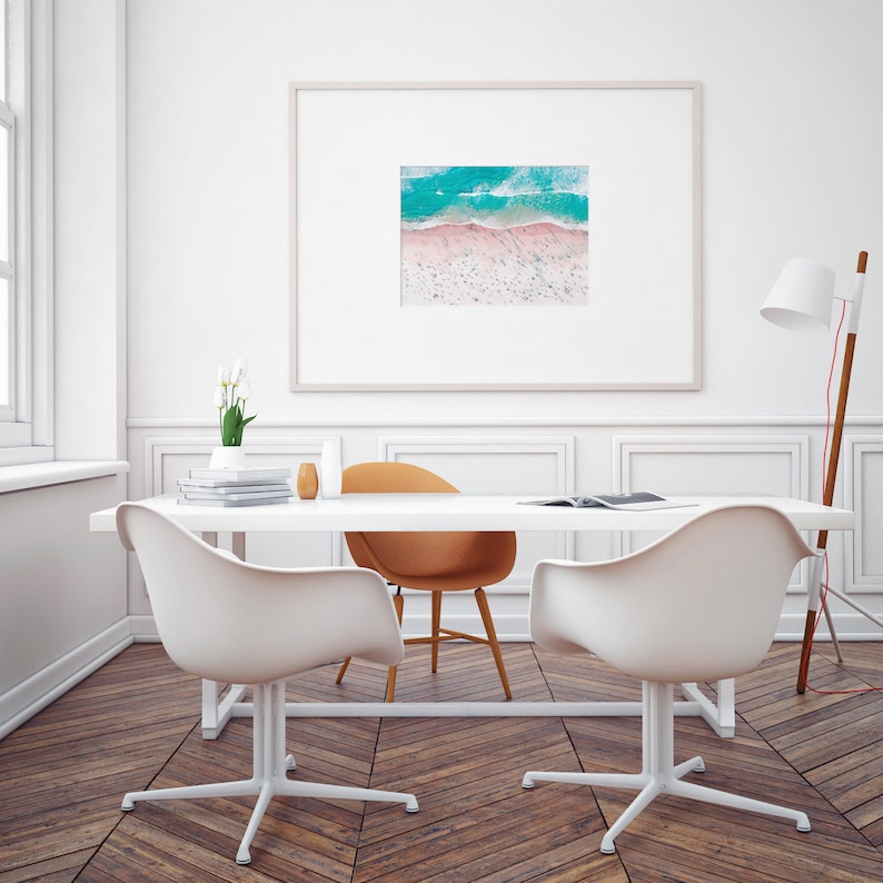 Op de afbeelding: Een wit bureau met een ingelijste foto van een luchtfoto van een strand met turquoise water en roze zand. Twee witte stoelen en een bruine stoel staan rond het bureau. Een lamp met een witte kap staat op de achtergrond.