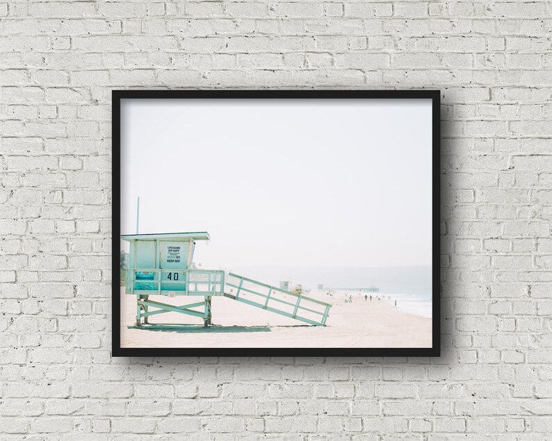 May include: A light blue lifeguard stand with a white roof and a sign that reads "Lifeguard Station 40 Keep Out". The stand is on a sandy beach with a pier in the distance.