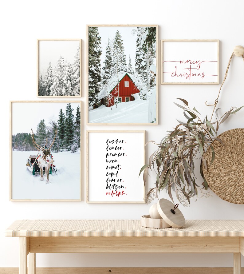 Op de afbeelding: Een set van vijf ingelijste prints met een winterthema. De prints tonen een rode hut in een besneeuwd bos, een rendier dat een slee trekt en een lijst met rendiernamen. De prints zijn gerangschikt op een witte muur met een houten bank eronder. De tekst op de prints luidt "Jester, Dancer, Prancer, Vixen, Comet, Cupid, Donner, Blitzen, Rudolph" en "merry christmas".