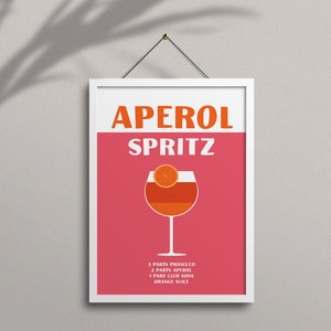 Pode incluir: Um p&ocirc;ster emoldurado branco com fundo rosa e o texto "Aperol Spritz" em laranja e branco. Um copo de Aperol Spritz com uma fatia de laranja &eacute; mostrado no centro. Os ingredientes s&atilde;o listados abaixo do copo: 3 partes de Prosecco, 2 partes de Aperol, 1 parte de &aacute;gua com g&aacute;s, fatia de laranja.